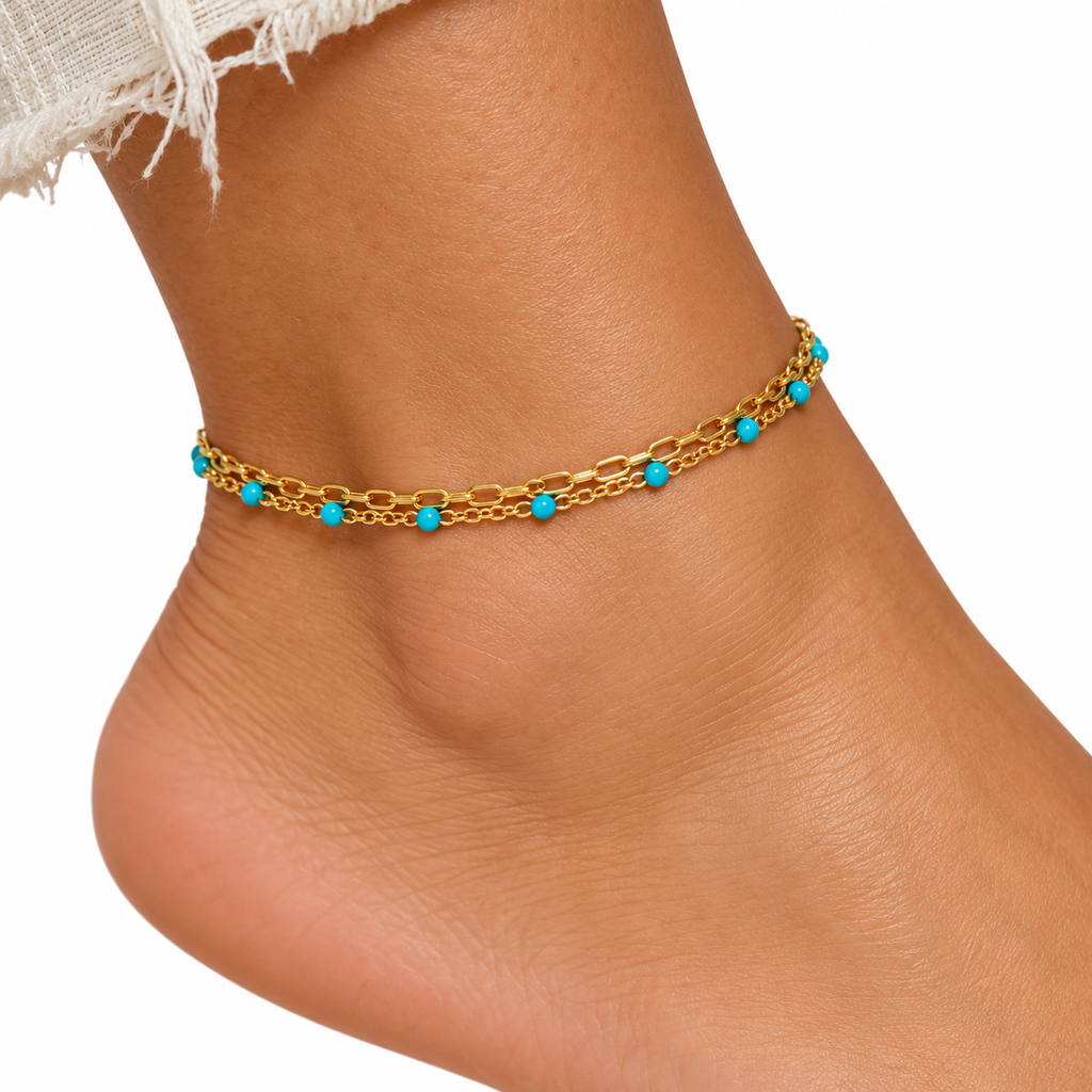 A delicate gold chain anklet with small turquoise blue bead accents, shown worn on a person's ankle against a white background.